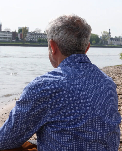 Reinhard Allgaier sitzt mit dem Rücken zur Kamera am Rheinufer und schaut aufs Wasser – Stille und Rückzug beim Achtsamkeitstag