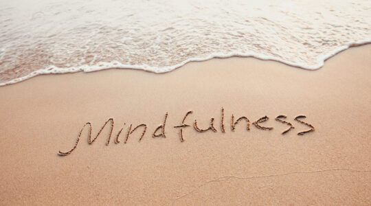 Das Wort „Mindfulness“ in Sand geschrieben, eine Welle läuft heran