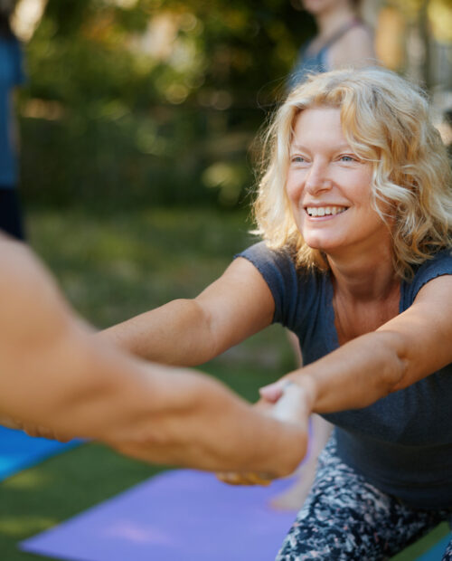 Zwei Frauen halten sich lachend an den Händen und dehnen sich gemeinsam auf Yogamatten im Freien