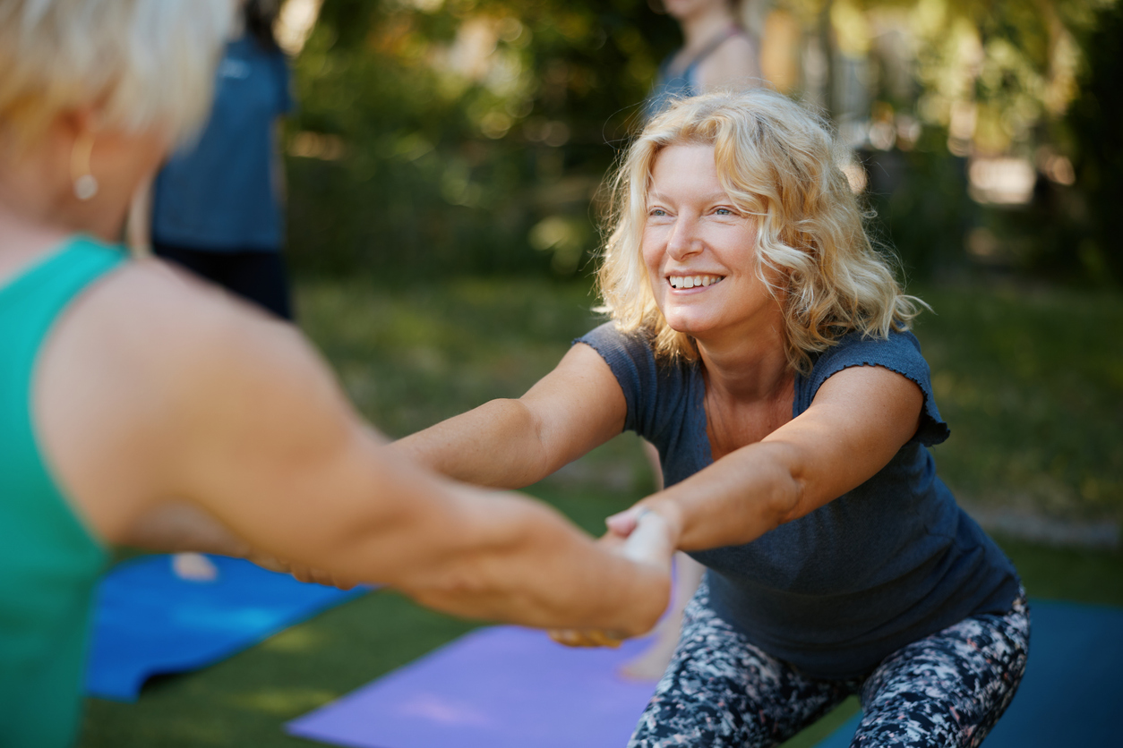 Zwei Frauen halten sich lachend an den Händen und dehnen sich gemeinsam auf Yogamatten im Freien